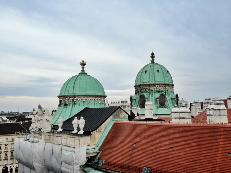 Panoramic view of Vienna with two prominent green-domed buildings rising above the historic cityscape under a cloudy sky.